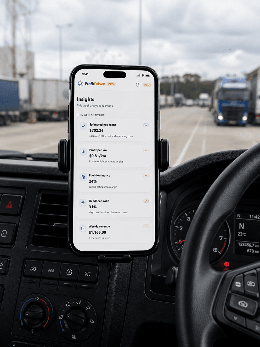 Smartphone mounted on truck dashboard displaying profit analytics app with semi-trucks in the background.